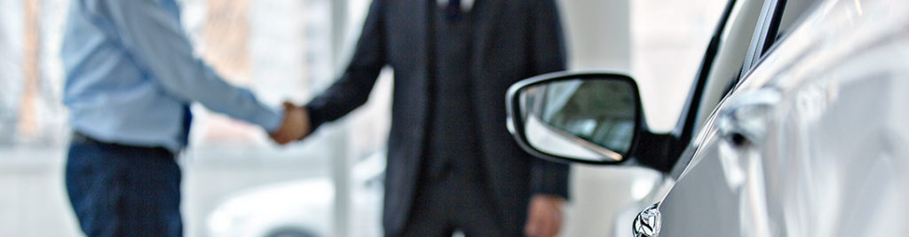 Two people in business attire shake hands in a blurred background, with a focus on the side mirror and part of a car in the foreground.