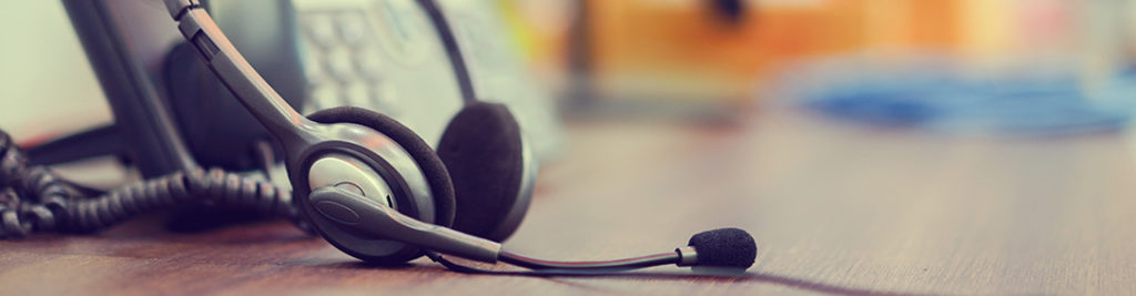 A headset with a microphone rests on a wooden surface in front of a blurred office phone. The scene suggests a call center or office environment.