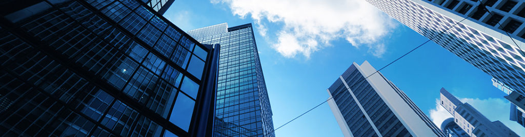 A low-angle view of several skyscrapers against a bright blue sky with scattered clouds. The buildings, featuring modern glass and steel architecture, create a dynamic cityscape. Power lines intersect the scene, adding an urban element.