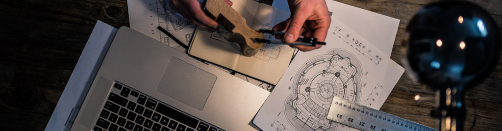 Overhead view of a desk with a laptop, architectural blueprints, a wooden model, and hands holding a caliper. A desk lamp illuminates the workspace, highlighting the detailed sketches and plans.