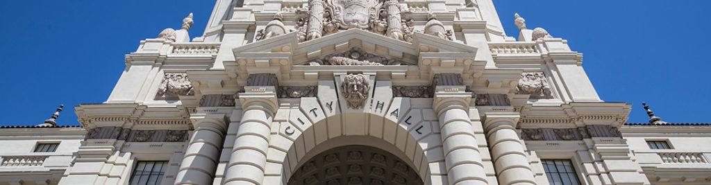 A low-angle view of an ornate city hall building featuring intricate architectural details, columns, and sculptures against a clear blue sky. The words "City Hall" are prominently displayed above the entrance.