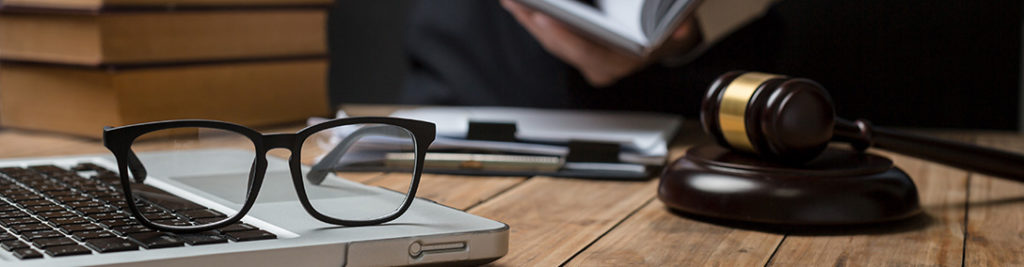 A pair of glasses rests on a laptop keyboard next to a gavel on a wooden desk. In the background, a person reads a book. The scene suggests a legal or academic setting.