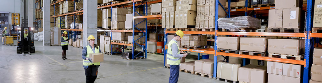 A warehouse scene with workers in safety vests and helmets organizing boxes on high shelves. One worker carries a box, another uses a tablet, and a forklift operates in the background. Shelves are filled with cardboard boxes and wrapped pallets.