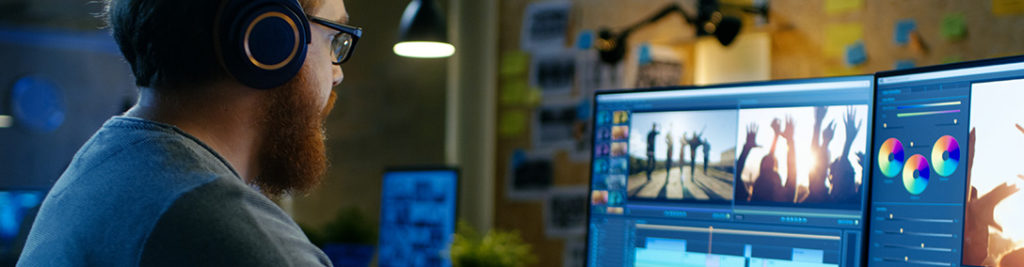 A person wearing headphones is sitting at a desk editing video on dual monitors. The left monitor shows a video timeline, while the right displays video clips and color grading wheels. The room is dimly lit with a creative workspace vibe.