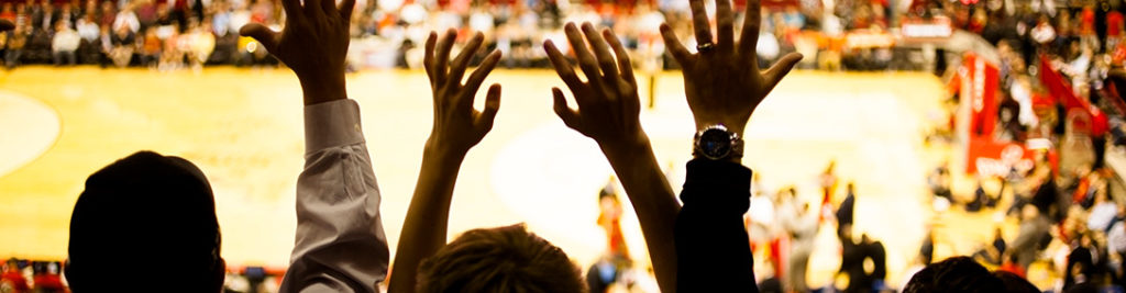 Three silhouetted individuals raise their hands in excitement, watching a basketball game in a crowded arena. The background shows a blurry basketball court with players and spectators in the stands.