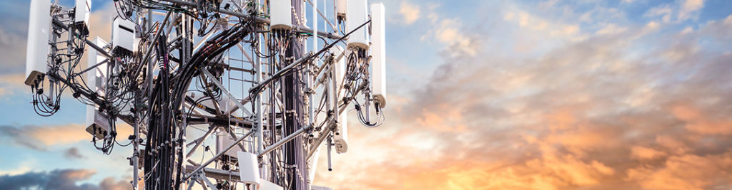 A telecommunications tower with numerous antennas and cables stands against a vibrant sky at sunset, showcasing a blend of orange and blue hues.