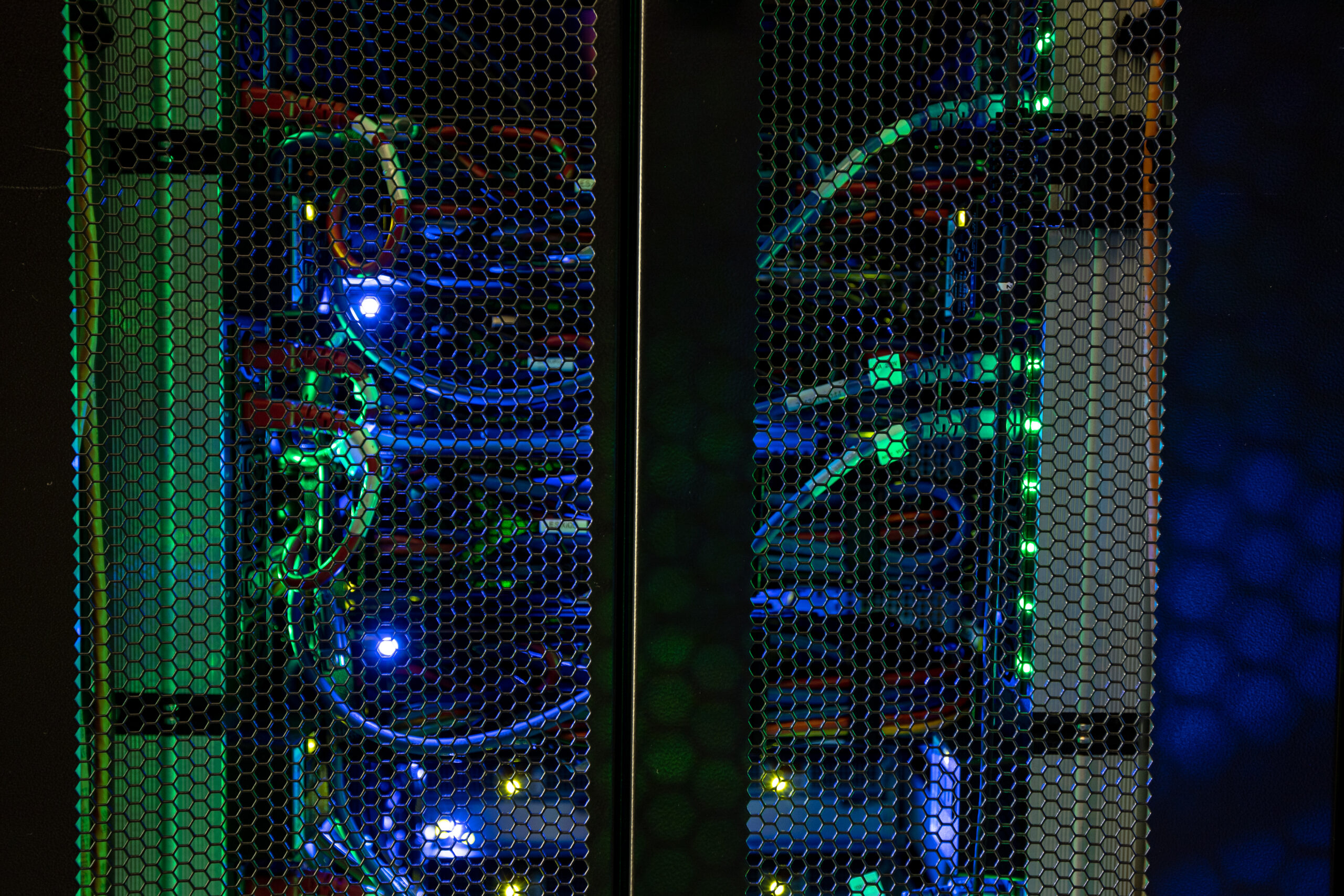 A dimly lit server rack with glowing blue and green LED lights, cables, and network equipment visible through a perforated metal door.