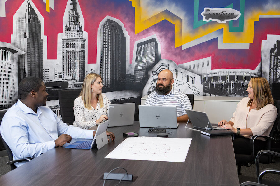 Four people sit around a conference table with laptops during a meeting. A colorful mural of city buildings and abstract shapes adorns the wall behind them. They appear engaged in conversation.