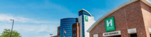 Brick building with "West Entrance" sign and a logo with an "H" in a green box. Adjacent to a modern blue-glass structure. Bright blue sky and trees in the background.